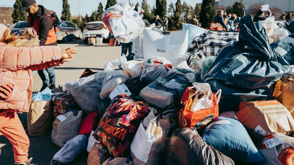 A man with long blond hair and a full red beard, wearing a dark blue beanie and a black worn and weathered jacket, is outdoors at a waste disposal or recycling site. He is standing beside a large, rus