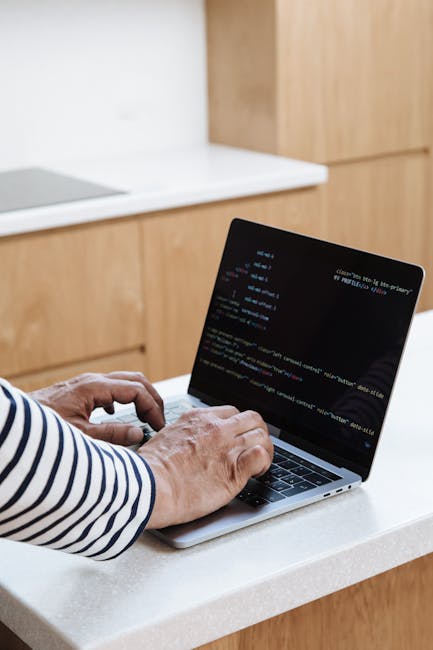Close-up of a person’s hand typing on a silver laptop keyboard placed on a wooden desk. The laptop screen displays lines of code with a dark background and brightly colored syntax. The individual is