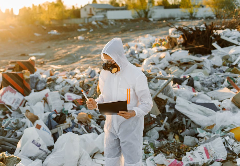 A person wearing a white hazmat suit, protective goggles, and a black respirator mask stands in a landfill or dump site filled with scattered waste and rubbish. They are holding a clipboard or tablet