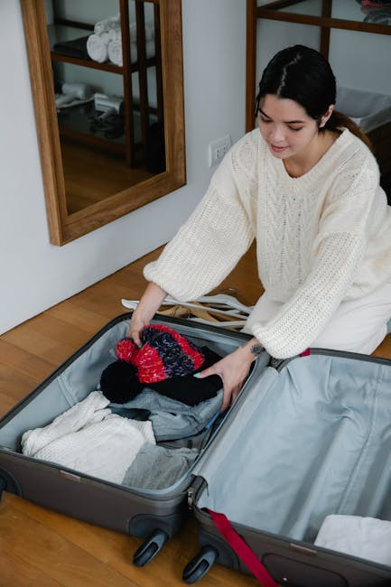 A young woman with short black hair, dressed in a cream-colored knitted sweater, is kneeling on a wooden floor as she loads clothing and accessories into an open, large grey suitcase with red accents.