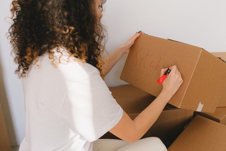 A young woman with short black hair, dressed in a cream-colored knitted sweater, is kneeling on a wooden floor as she loads clothing and accessories into an open, large grey suitcase with red accents.
