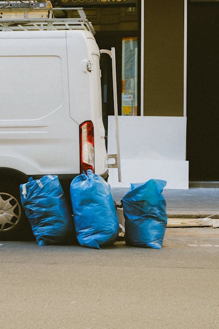 A white commercial van parked on a city street with its rear doors partially visible, positioned next to three large blue rubbish bags made of thick plastic, which are placed on the pavement close to 